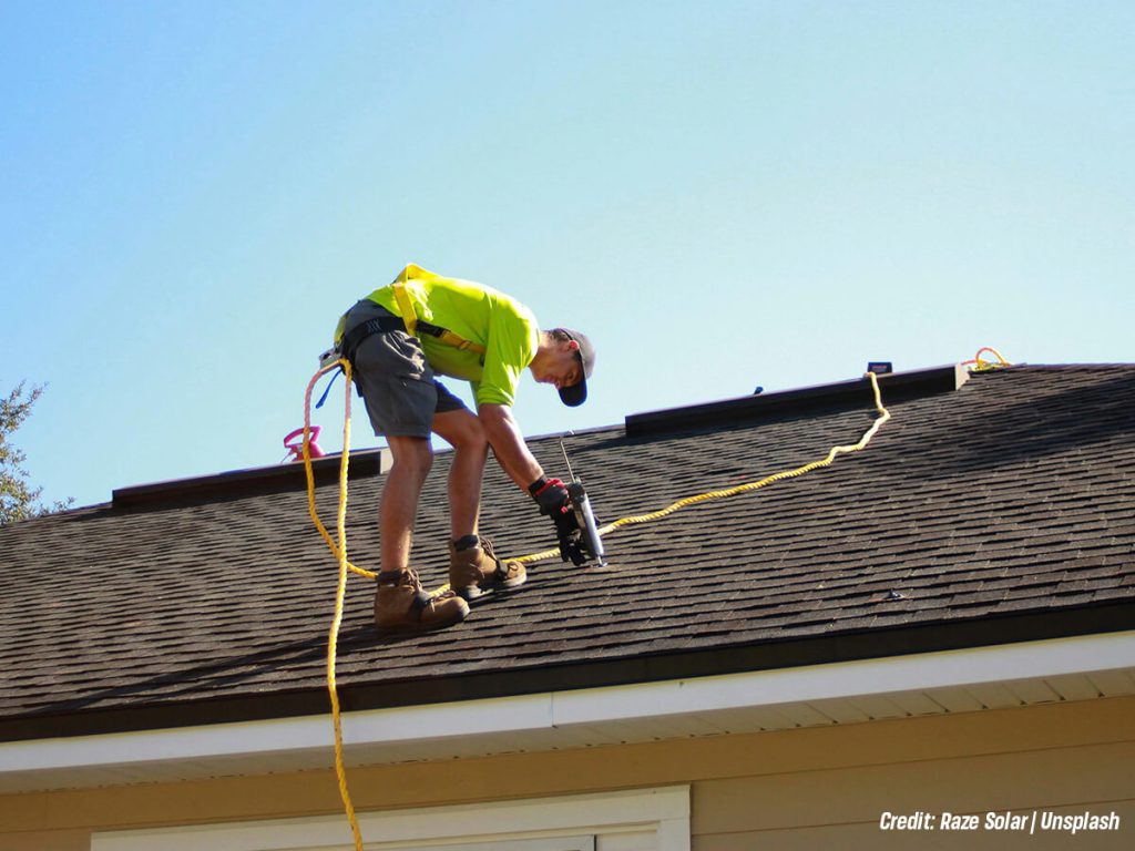Roofing worker on a pitched roof secures a yellow safety rope, wearing a bright lime shirt, shorts, and boots with a tool in hand.