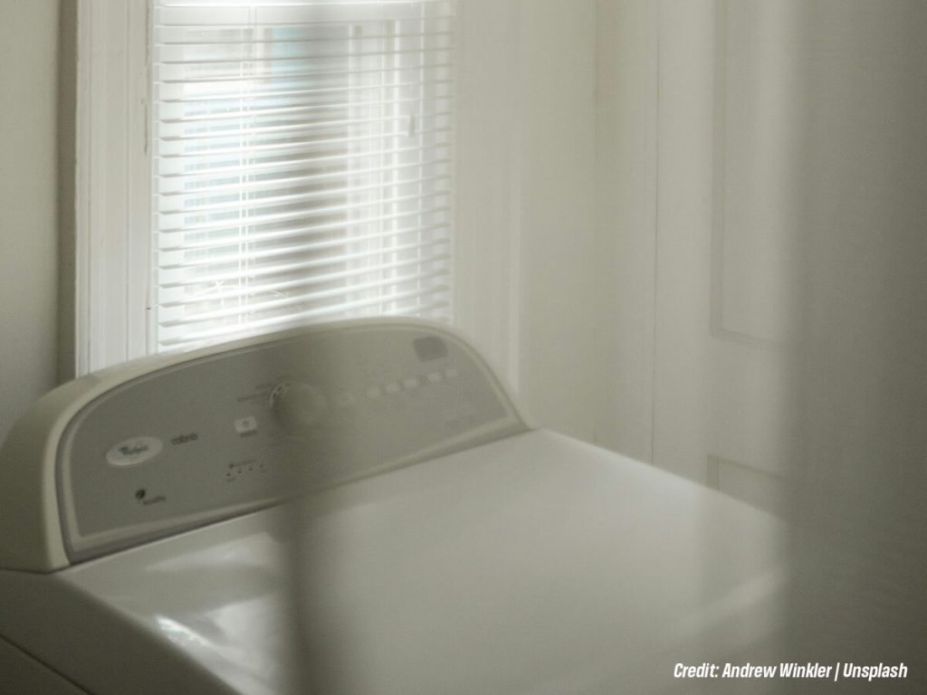 Top-loading washing machine in a bright laundry room beside a window with horizontal blinds.