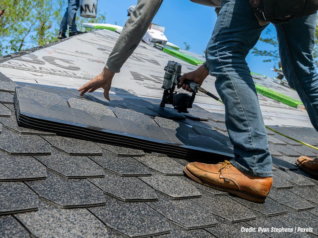 Roofer installs dark asphalt shingles on a sloped roof using a nail gun and stacking shingles nearby on the roof surface, with boots visible.